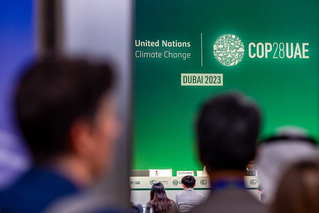 Attendees entering a meeting room during COP28, looking at the main stage with a green backdrop stating: “United Nations Climate Change | COP28UAE; Dubai 2023.”