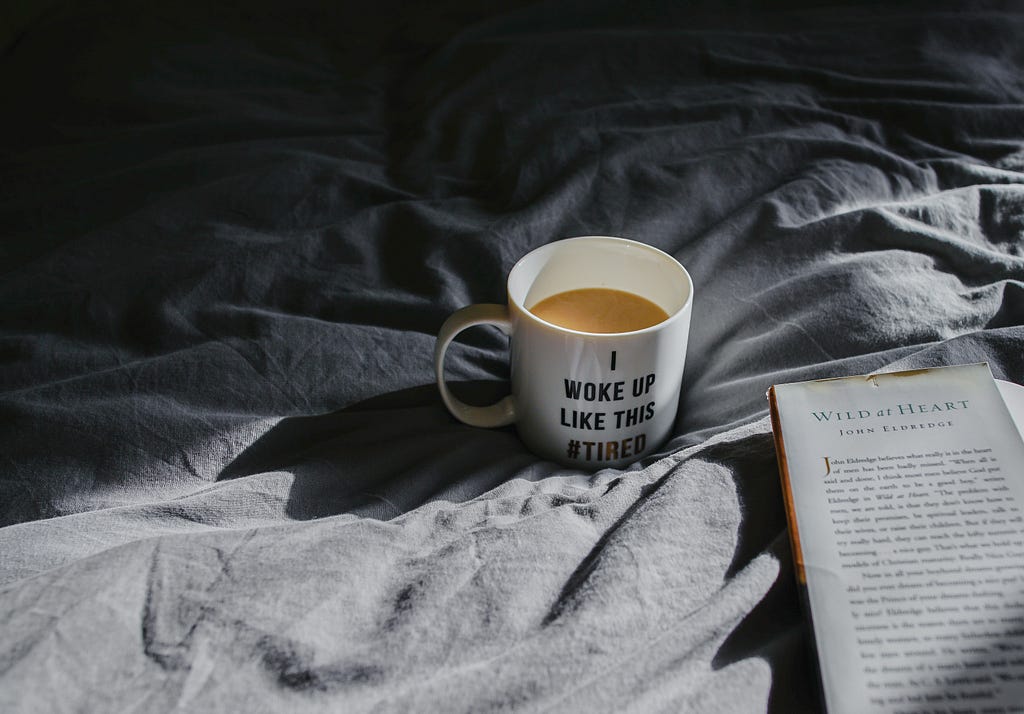 photo of a coffee mug that reads “I woke up like this #tired.” The mug is sitting on gray sheets beside an open book.