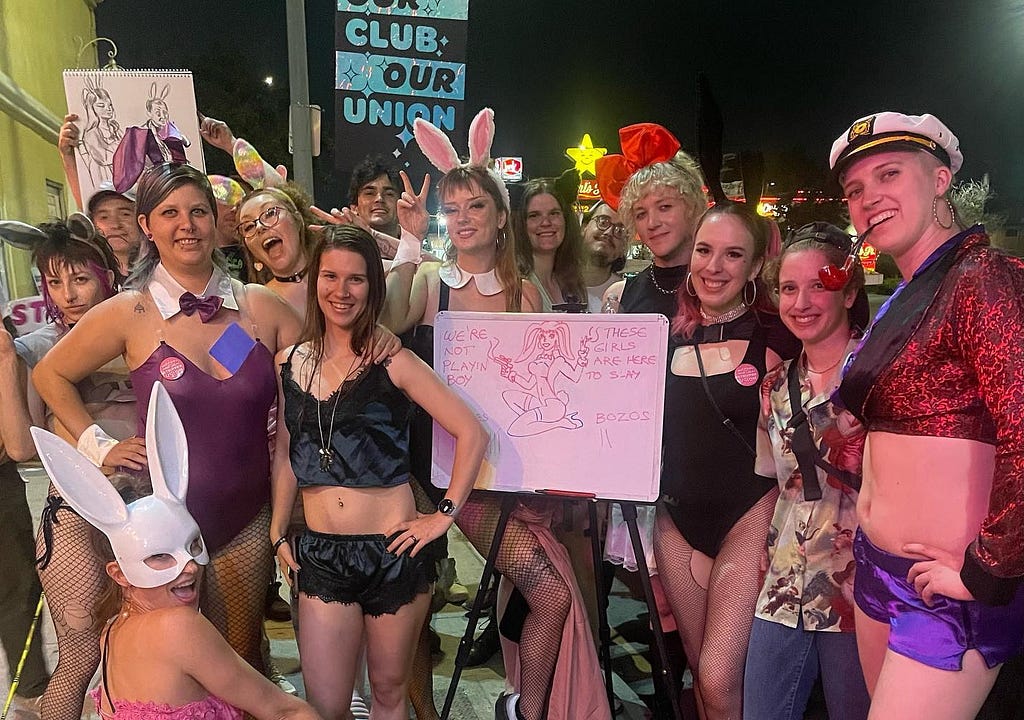Stripper-organizers from the Star Garden Topless bar celebrate a successful picket night. Many are dressed with a “playboy” costume theme — several are bunnies, and one is Hugh Hefner. The photo includes a whiteboard that tallies how many customers crossed the picket and how many were turned away, showing that the majority were turned away
