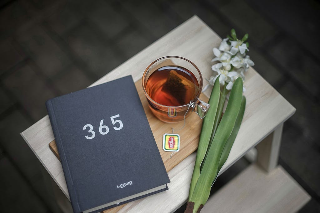 a book with three hundred sixty five on its cover next to a cup of tea and a stalk of lily of the valley