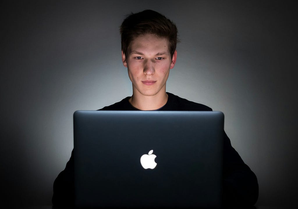 A young man is lit by the glow of his laptop in the dark.