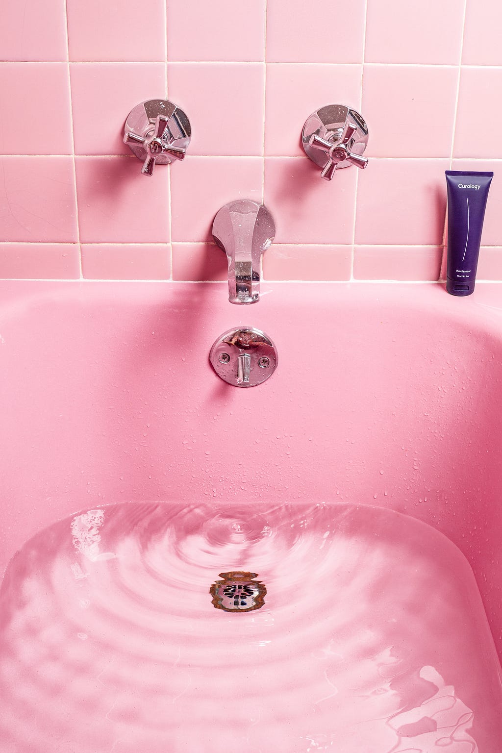 Pink bath tub filled with rippling clear water. Pink bathroom tile and silver faucet. Dark purple tube of Curology “the Cleanser” body care product on the right side of the tub faucet.