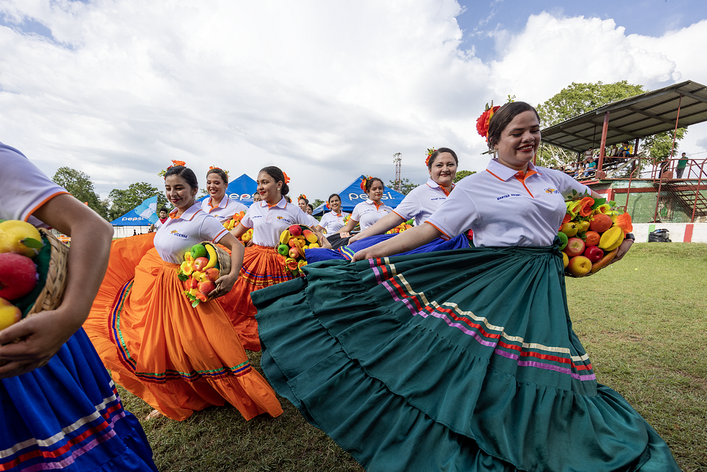 A group of women dance in formation while wearing colorful and sweeping long skirts and holding baskets of fruit.