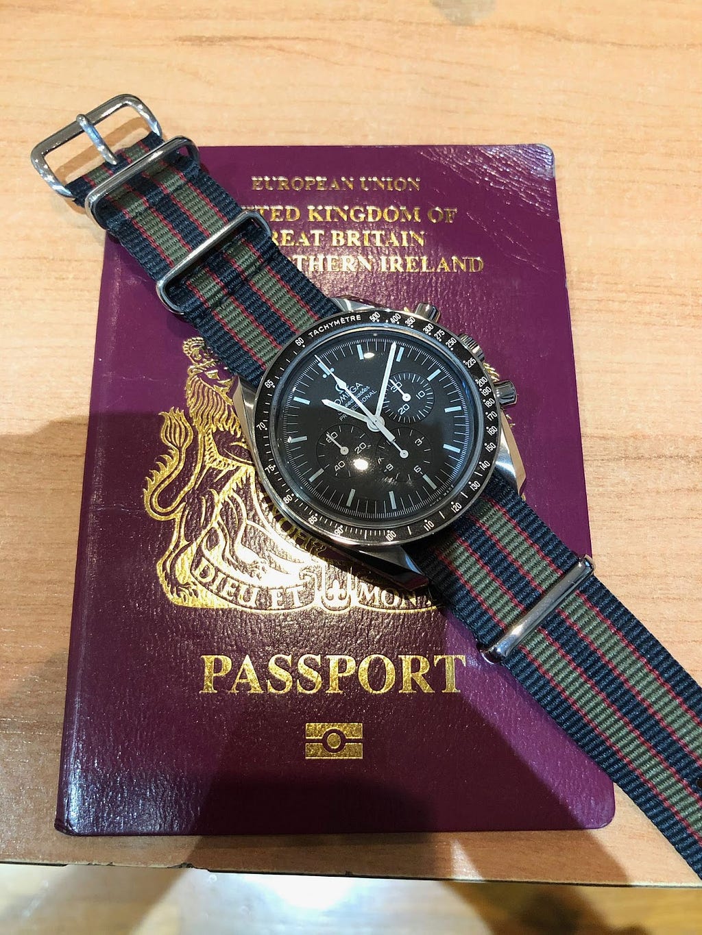 Older burgundy colour EU UK passport with an Omega Speedmaster Professional watch on a NATO strap cross it diagonally from top left. Strap back, red and green.