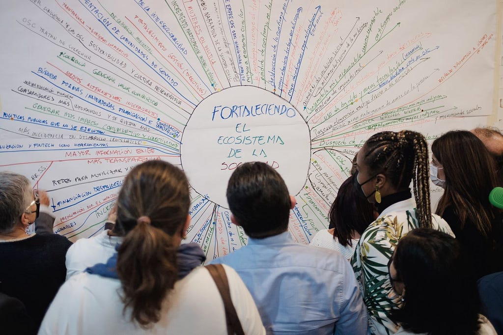 A group of people look at visual representation of their organization’s goals made to look like the sun, with a large circle at the center and dozens of lines with written text above them emerging from the circle as if like sunbeams.