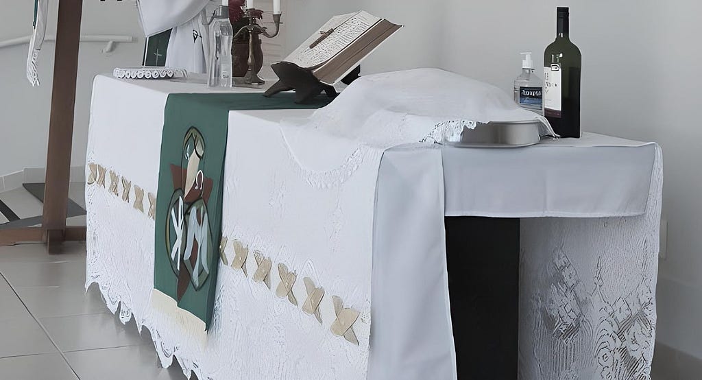 Mesa de igreja coberta com panos brancos e verdes. Em cima da mesa, vê-se uma bíblia aberta, uma garrafa de vinho e um vidro de álcool gel. Ao fundo, observa-se uma parte do tronco de um homem vestido com a batina religiosa.