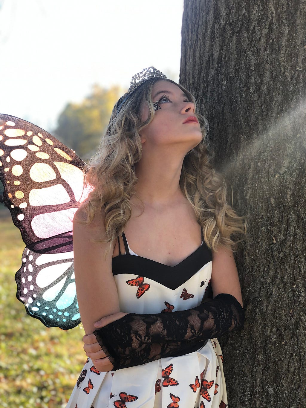 Young woman with long, curly blonde hair, wearing black and white butterfly wings in the style of a monarch butterfly, a silver tiara, and a white dress with orange monarchs, standing against a tree trunk and looking upwards with a serious expression.