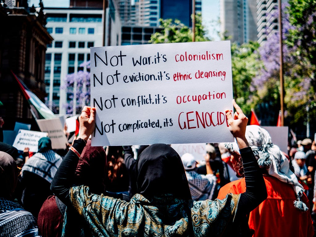 A protester in a crowd holds a sign that says, “Not war. It’s colonialism. Not eviction. It’s ethnic cleansing. Not conflict. It’s occupation. Not complicated. It’s genocide.”