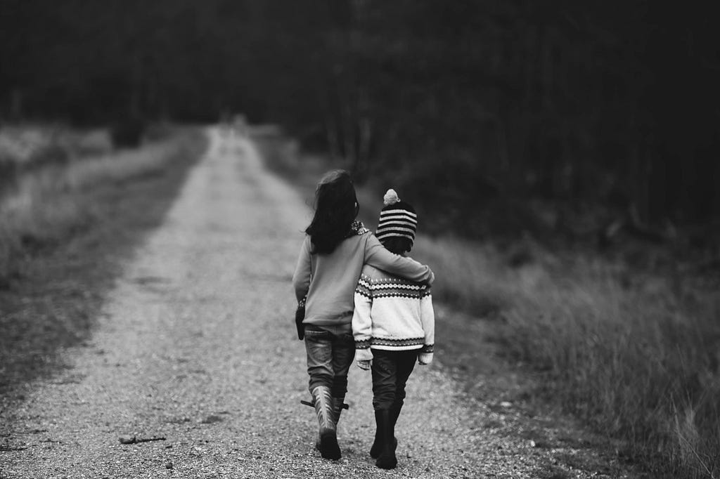 An black and white image shows two small children walking down a long road. The taller child has their hand around the shoulders of the smaller child. They have their backs to us.