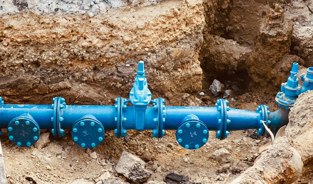 A blue pipe is buried in a trench. The pipe has four flanges with bolts. There is dirt and a large rock next to the pipe.