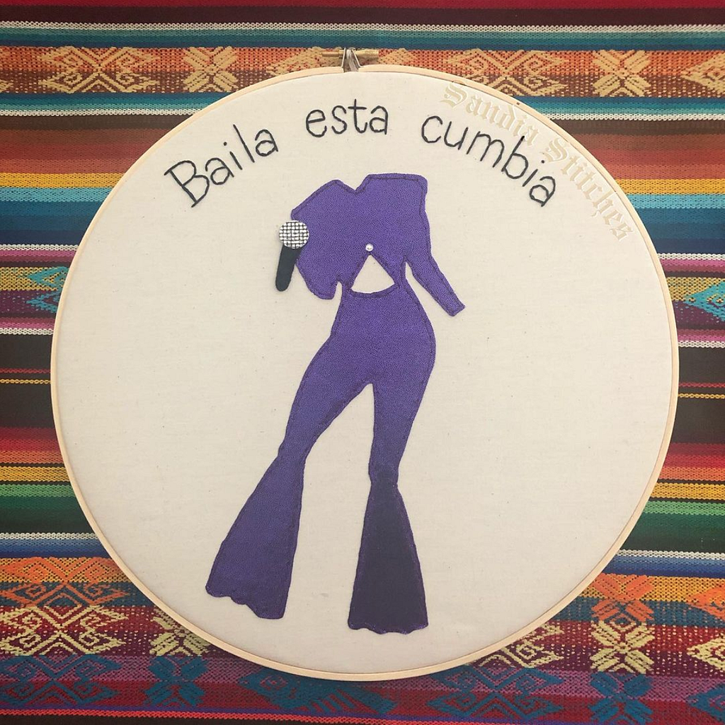 A circular embroidery hoop with the words “Baila esta cumbia” and the image of a purple jumpsuit with a microphone. The background is a colorful Mexican tapestry.