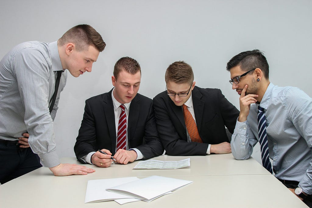 Four men looking at a paper on an office table. #AmWriting #Sarcasm #ShortStory