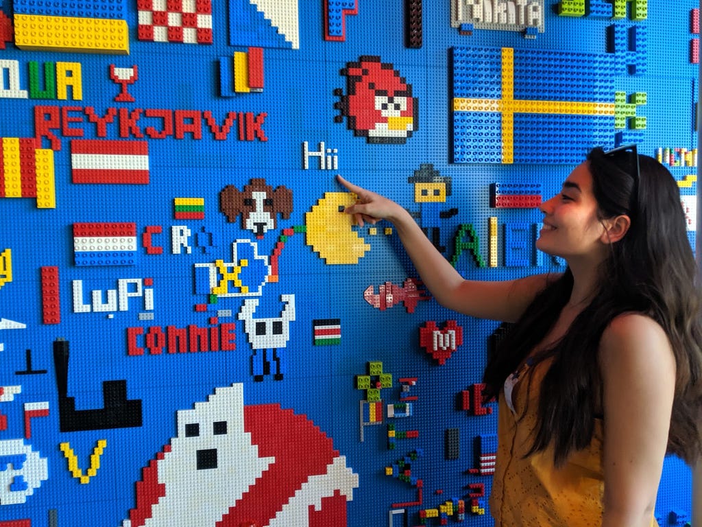 A person with long black hair smiles and points at a multi-colored wall made of legos.