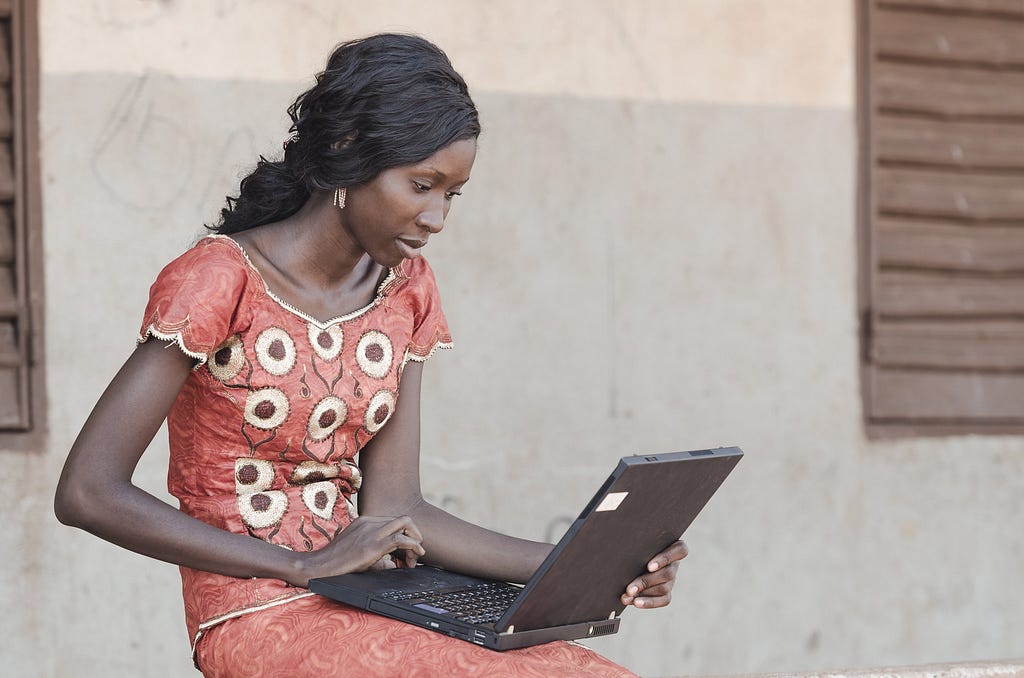 A woman sits outside of a building with stucco walls and wood shutters, working on a laptop.