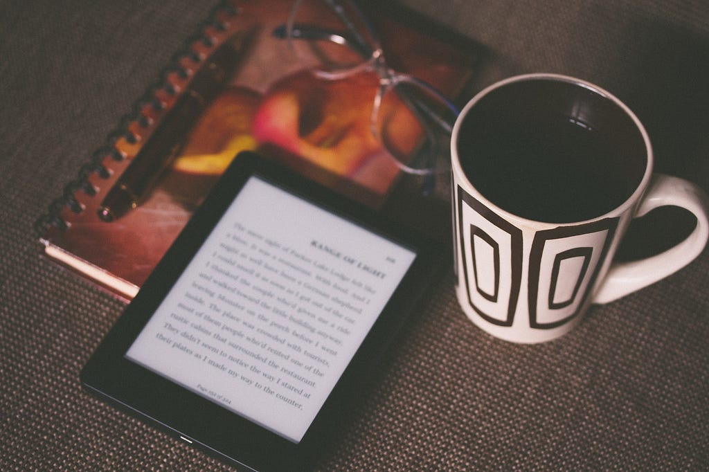 close up of kindle, coffee mug, notebook and glasses