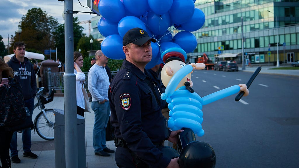 A city police officer holding a balloon police man toy while staring at the camera with a scowl.