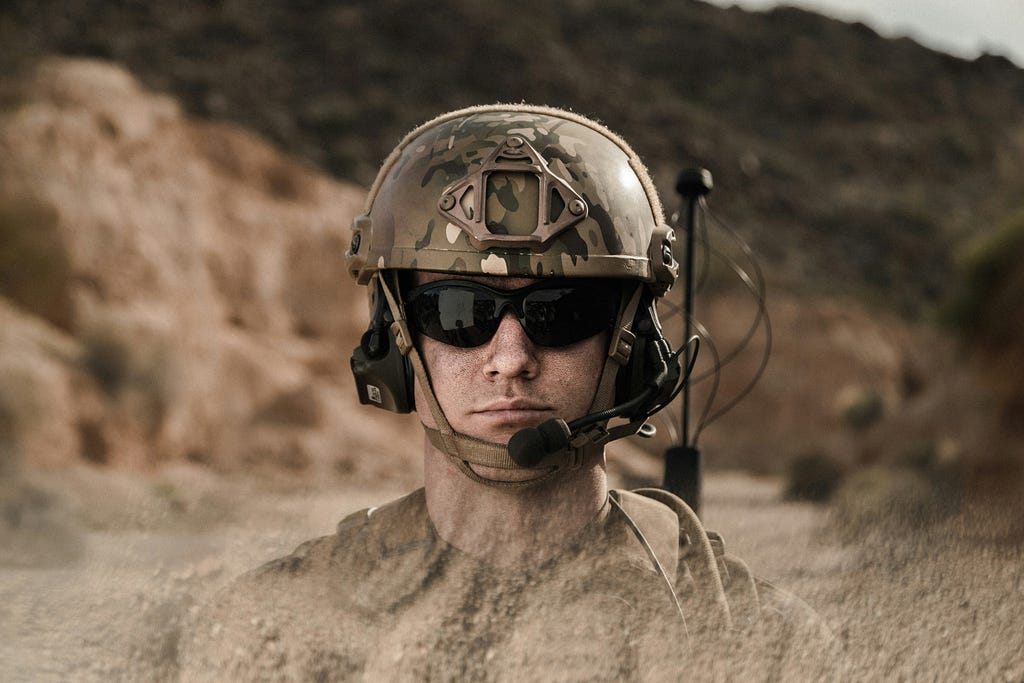The image of a soldier with helmets and glasses. He appears to be in a sandy desert like landscape.