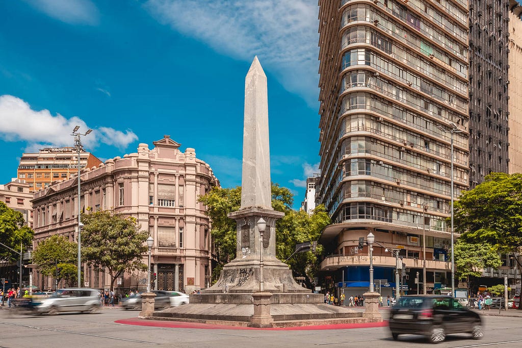 Foto do obelisco que fica em frente a praça 7 de setembro em BH. Em volta tem carros e prédios ao redor.