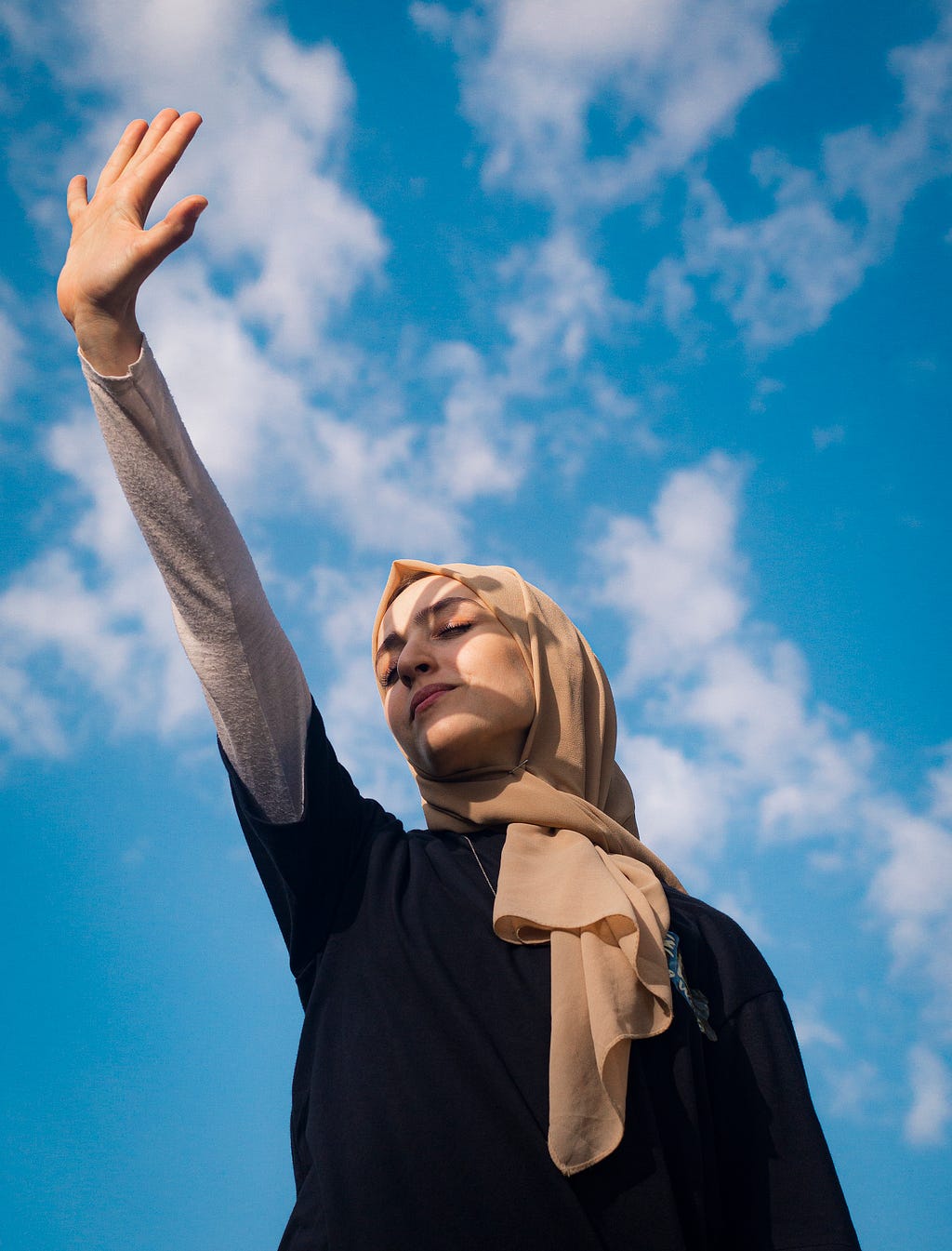 A woman raising her hand out to the sky with her eyes closed. Blue sky in the background.