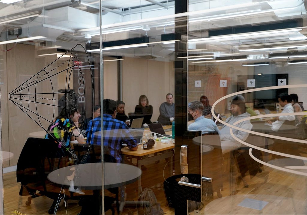 A behind-the-scenes image of our board of directors and advisors, staff, and community members at NYU ITP. The image is taken in front of a glass door. Inside the room, around 10 people gathered by a table, working on their laptops.