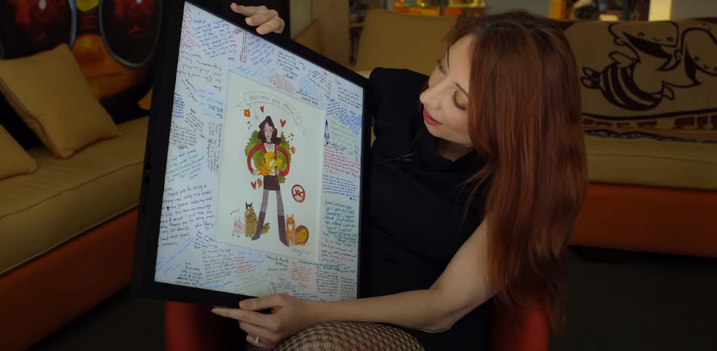 A resigned double fine veteran holds a large framed going-away card. At the center is a stylized drawing of her holding the Double Fine “two headed bee logo,” and flanked by cute animals. Along the border of the card, her various colleagues have left messages wishing her well in the next step of her journey