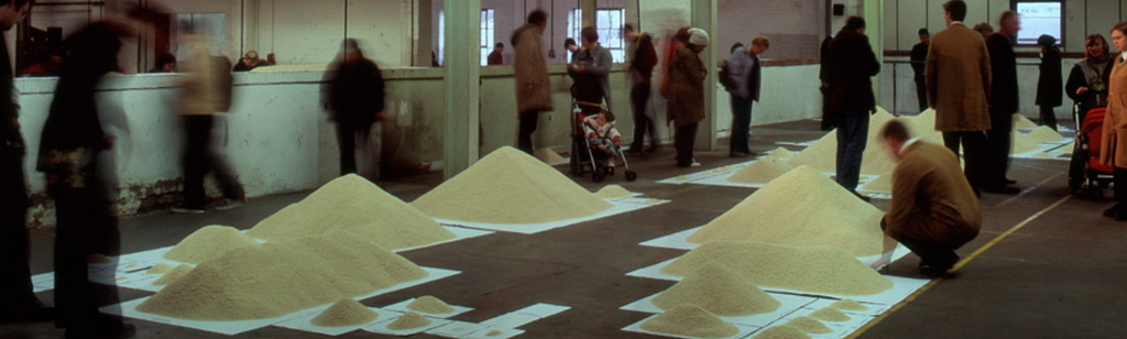 A large open space with an audience moving around the outskirts of a variety of sizes of piles of rice, carefully placed on top of correspondingly sized paper