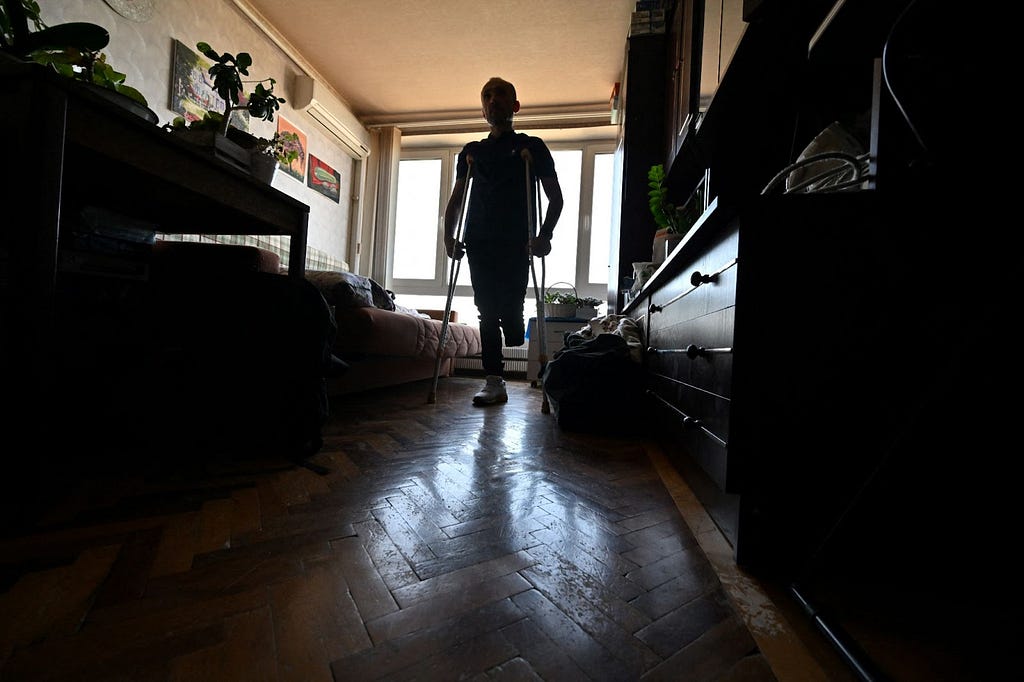 A silhouette of a man on crutches who is missing his left leg from the knee down as he stands in a small apartment with windows behind him.