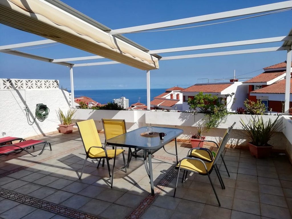 A sunny terrace with a table and four yellow chairs. There is a cup of tea on the table.