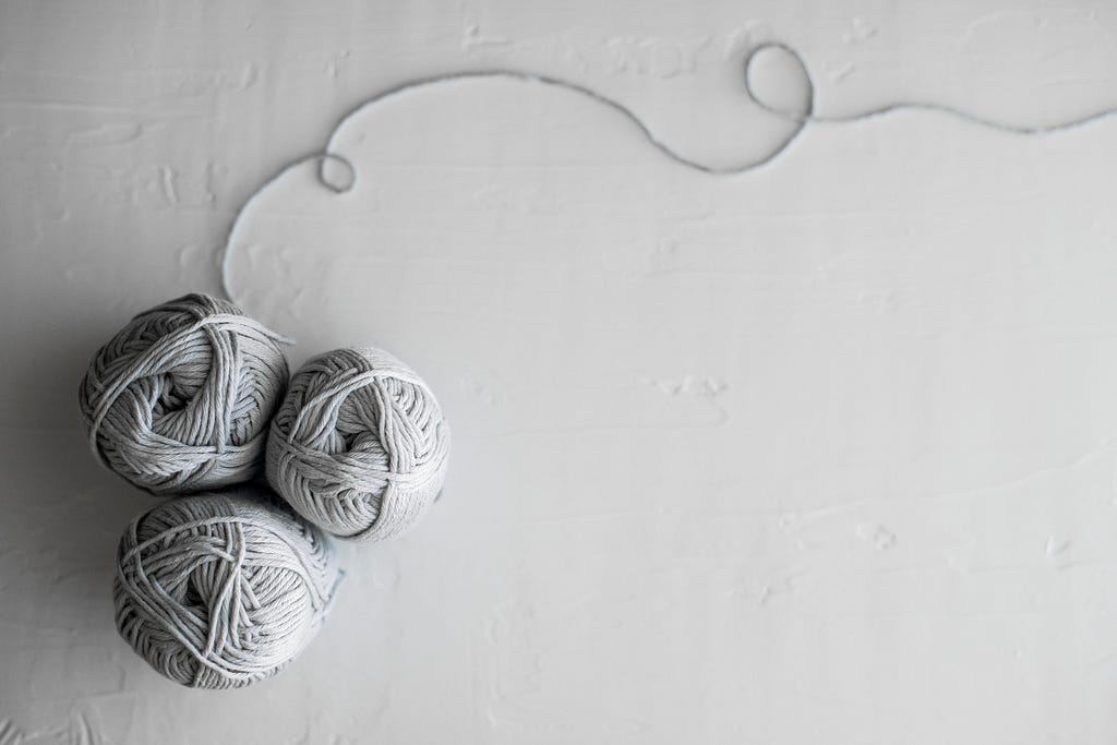 Three balls of light grey yarn with a singular thread looping at the top on a grey background