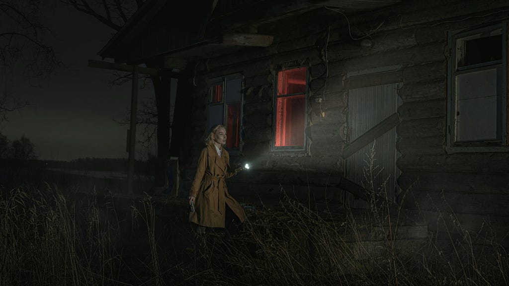 Woman outside of on old wooden house in the fields at night with a flashlight.