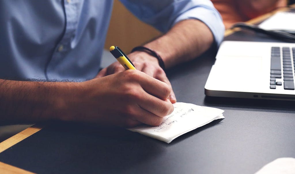 A man sitting at a desk, a close up of the edge of his laptop and a notepad