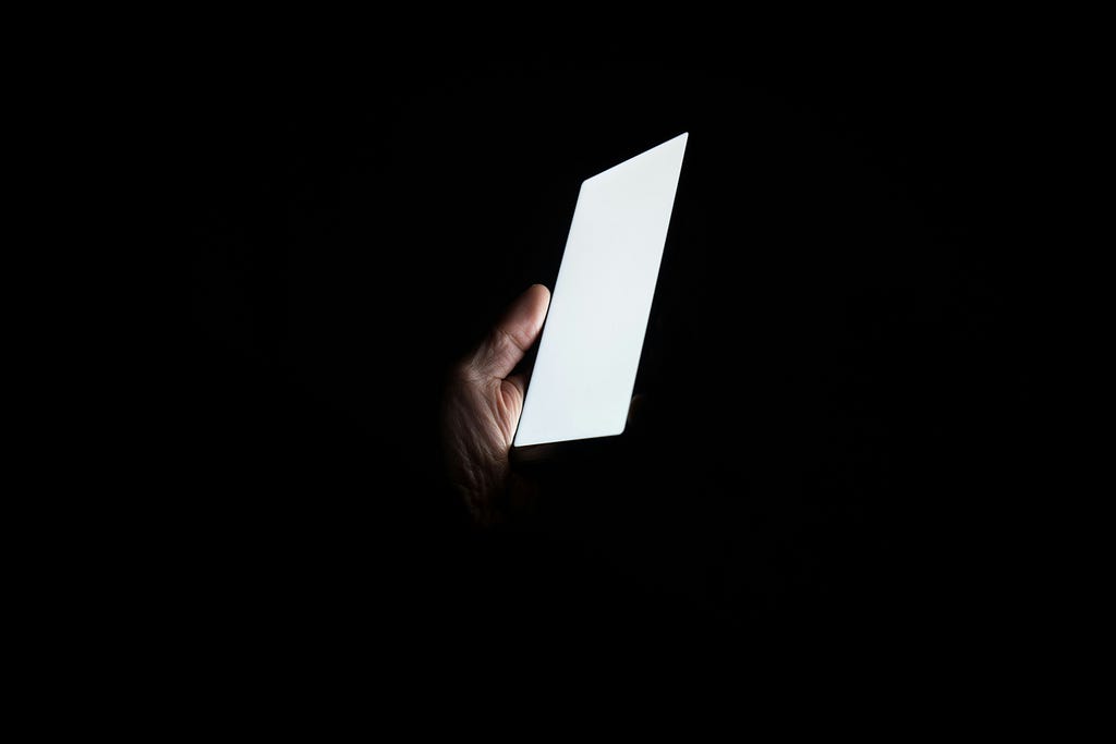 The white glow of a phone screen in a persons hand surrounded by complete darkness