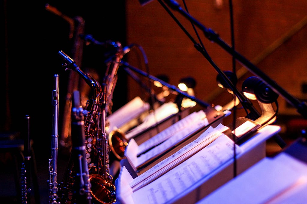 A image shows a jazz-like setup. There are 3 or 4 trumpets visible on the left. The right side has music sheets mounted on a stand. The background is of a room which is somber and is lit only by a golden light that falls aesthetically on the trumpets and music sheets.