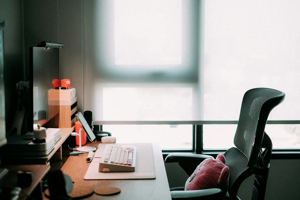A softly lit home office setup with a modern ergonomic chair, wooden desk, computer keyboard, and monitor. Warm light filters through a frosted window in the background, creating a calm, focused atmosphere. A pink cushion rests on the chair, adding a touch of comfort and personality to the workspace.