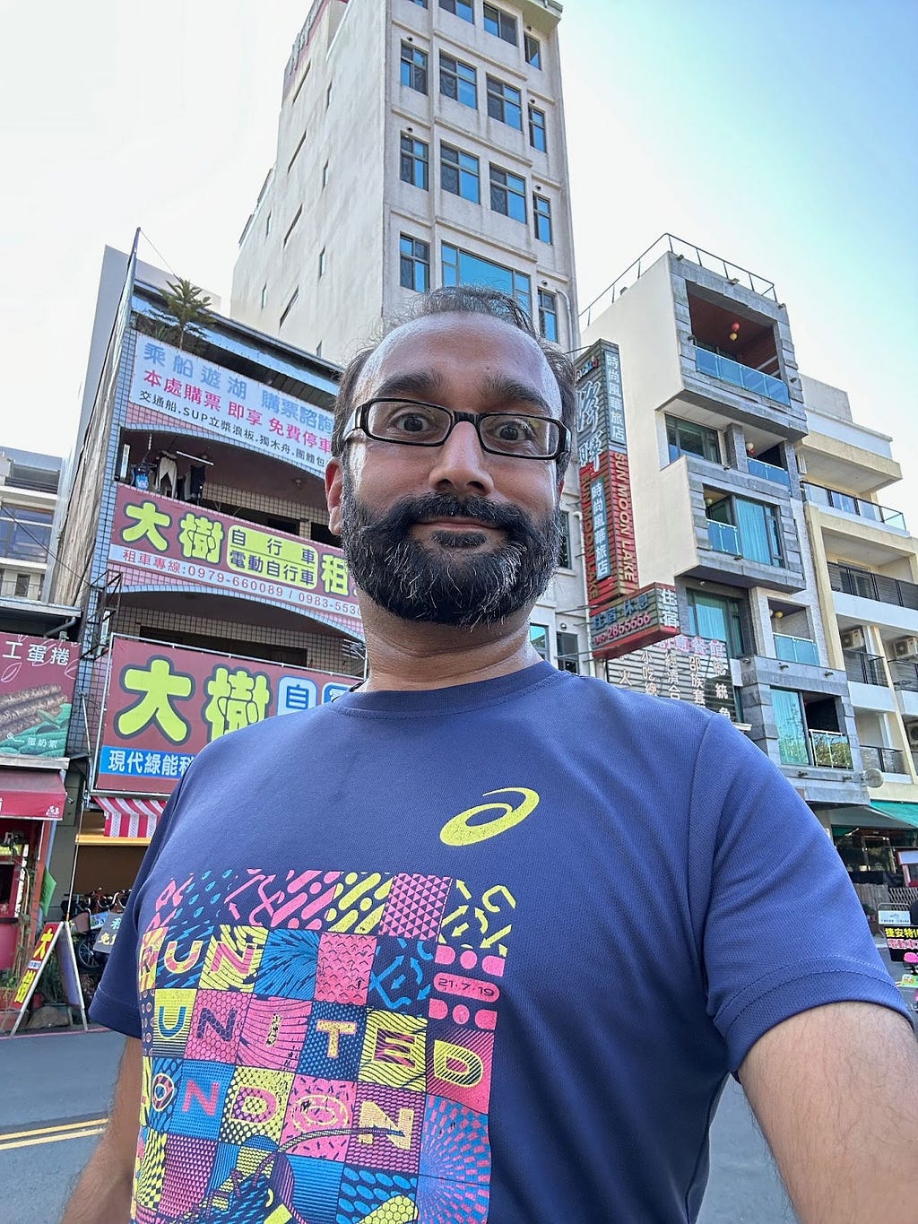 Selfie of brown man with beard and glasses in a dark blue running top with chinese shops and signs behind.
