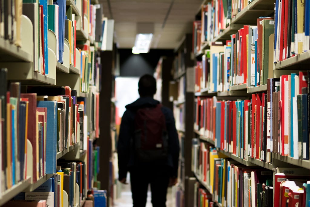 A person with a backpack standing between shelves in a library with their back turned.