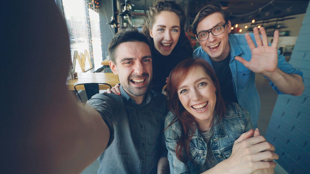 Point of view shot of cheerful group of young friends recording funny video in cafe posing and smiling hugging and gesturing. Friendship and happy people concept.