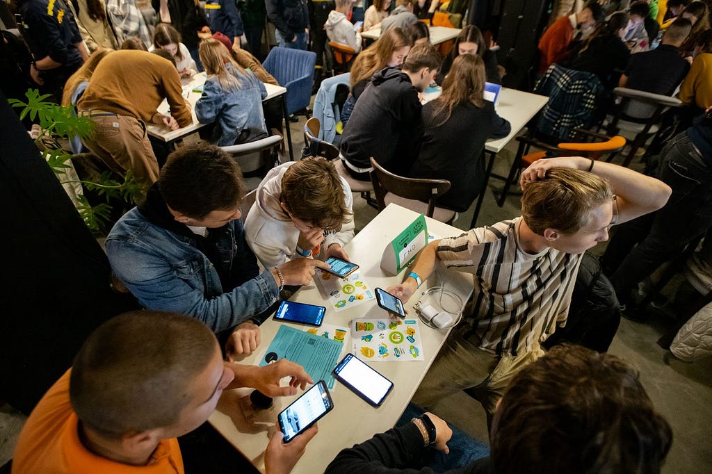 Groups of four to five young people sit at multiple tables holding mobile phones and reviewing paper materials that line their tables.