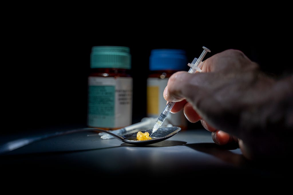 IV drug user uses needled syringe to draw up congealed drugs from a spoon with prescription bottles in the background.