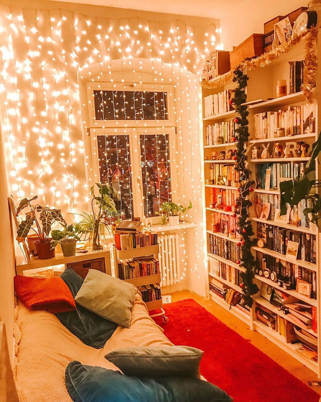 A reading room with a tall bookshelf full of books across from a comfy sofa with decorative pillows and fairy lights on the w