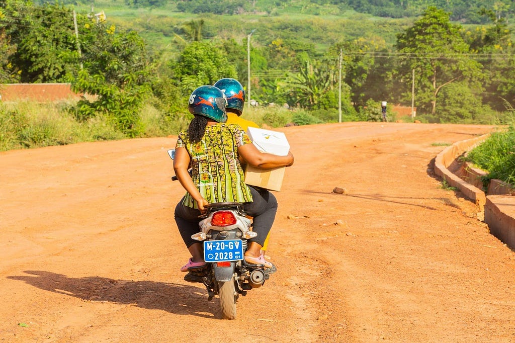 Judith on the back of a motorcycle, holding a box of medicine in her right arm and holding onto the motorcycle with her left hand.