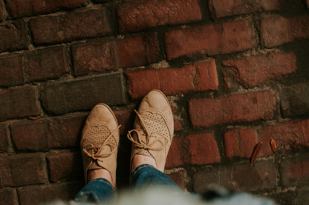 Photo of a brick floor with a person standing on it, the only thing in sight are a couple of cinamon colored leather shoes.