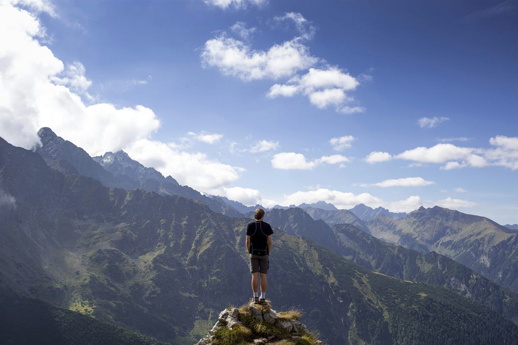 A man standing at the top of a mountain under a sunny sky. He’s free of his Imposter Syndrome