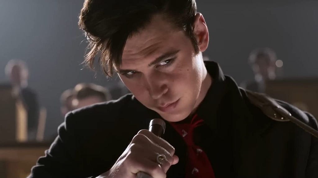 Austin Butler is pictured in his Elvis attire. He is wearing a guitar on a guitar strap and is holding the microphone in front of him.