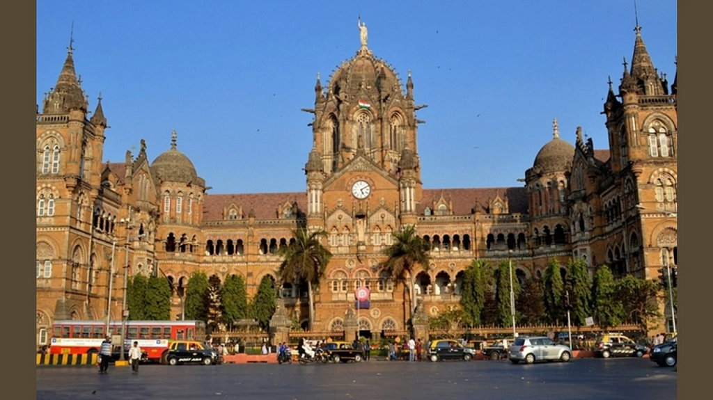 Chhatrapati Shivaji Maharaj Terminus in Mumbai, a UNESCO World Heritage Site. The grand Gothic-style railway station features domes, pointed arches, and a central clock tower with a statue on top. The foreground shows busy city traffic with taxis, buses, and pedestrians.