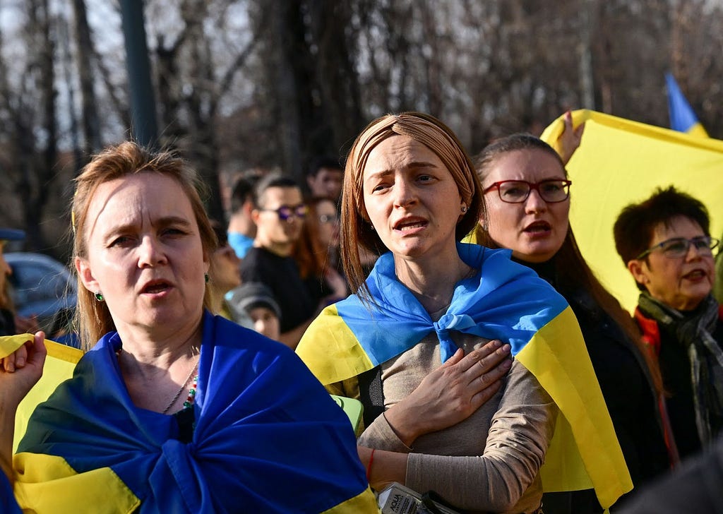 Two women draped in Ukraine’s yellow and blue flag sing while standing outside among a crowd of others protesting the war in Ukraine. One women raises her fist and the other woman holds her right hand over her heart.