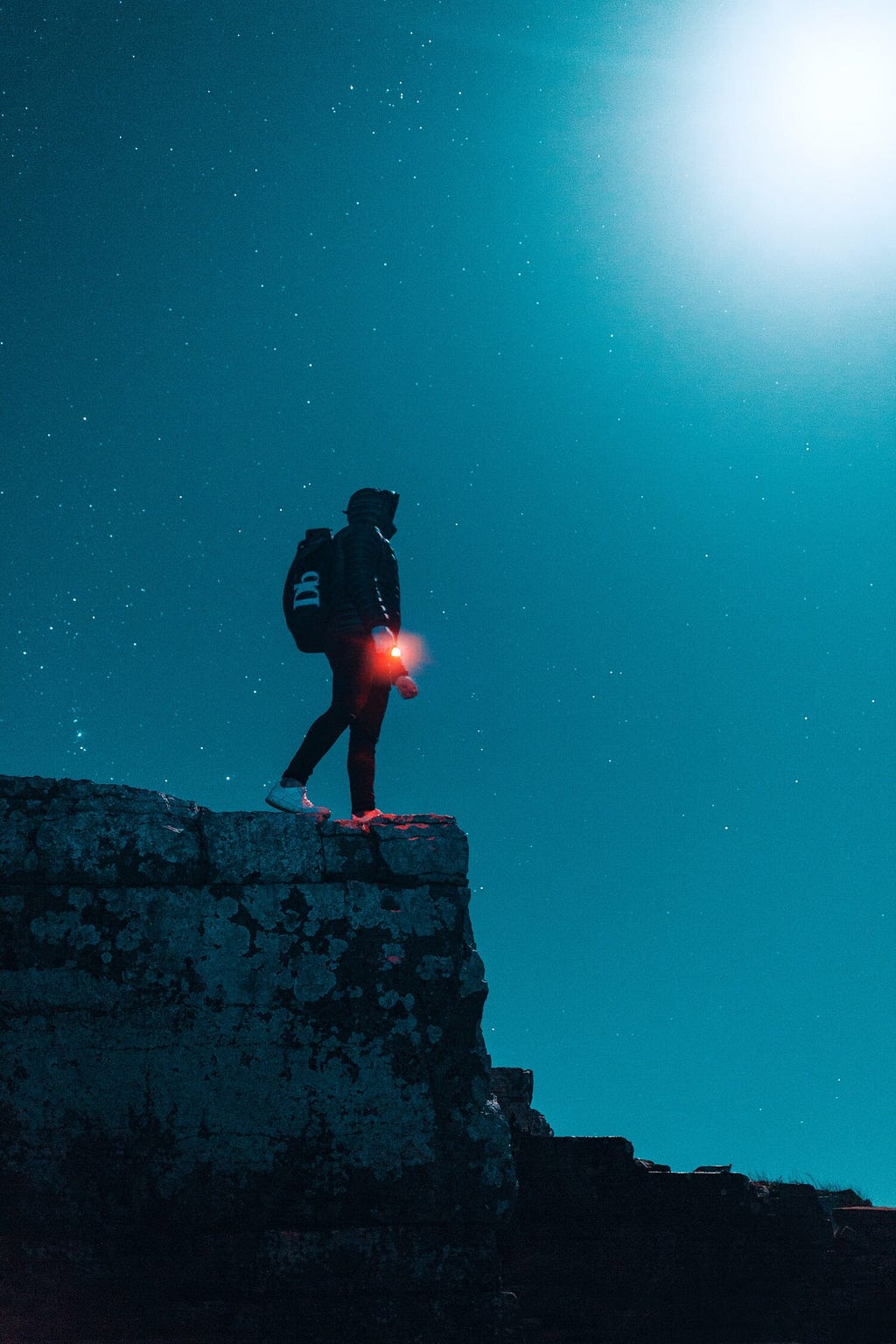 A man on a cliff looking at the night sky witha red glowing beacon in his hand