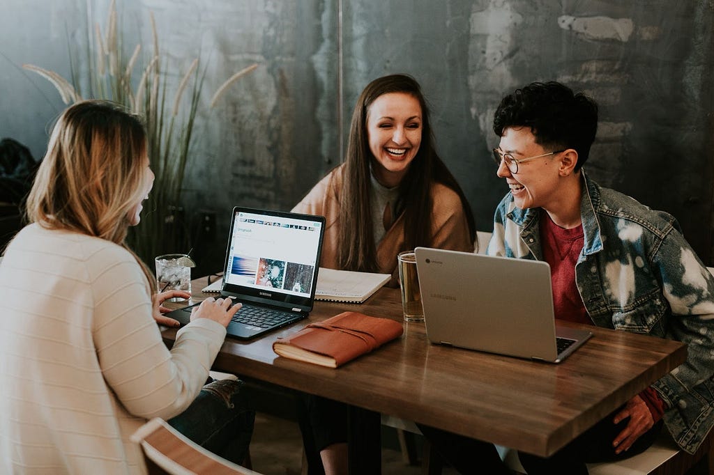 Three people sit at a table with laptops and have a conversation