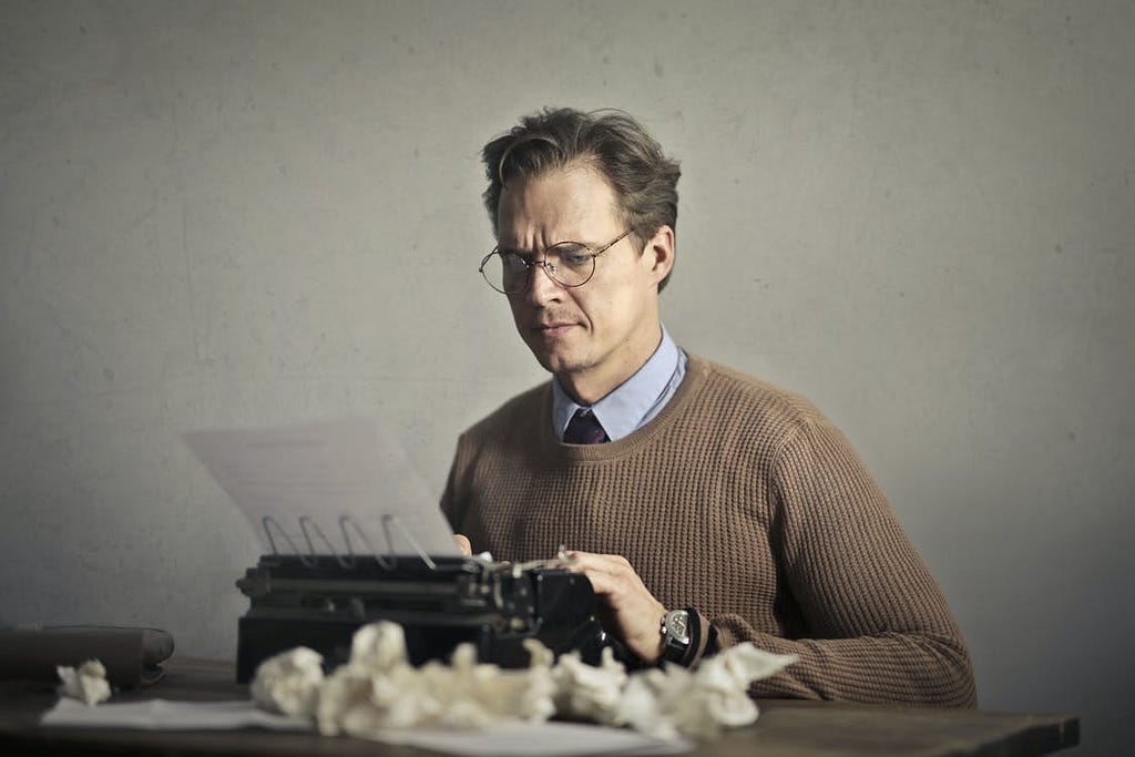 A frustrated-looking man writing a document on an antique typewriter with several crumpled up drafts on the table in front of him.