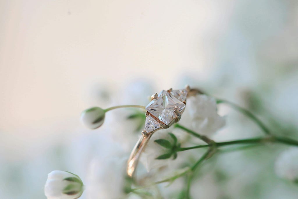 Close-up of a custom engagement ring featuring a stunning diamond centerpiece, surrounded by delicate white flowers. The elegant design of the engagement ring is highlighted against the soft, natural background.